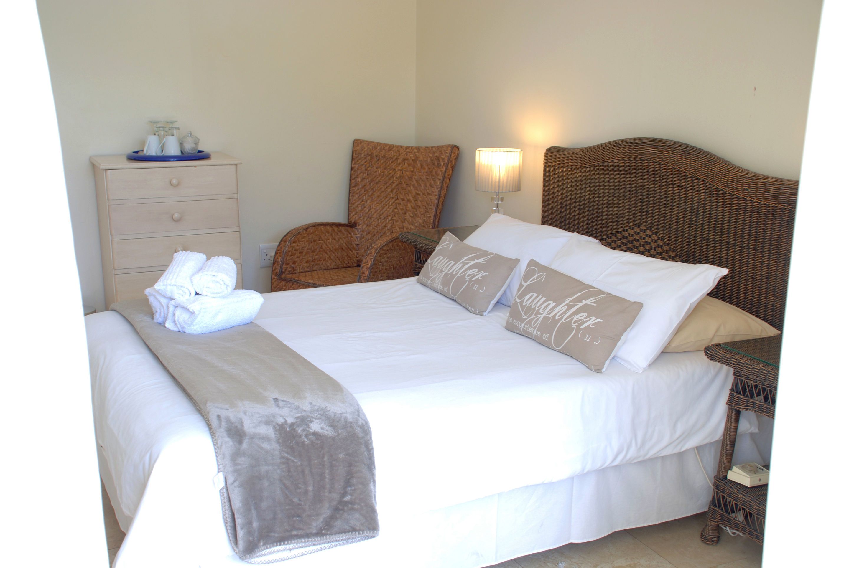 A neatly made bed with a white comforter and pillows, a wooden nightstand with a lamp, a wicker chair, and a dresser with folded towels.
