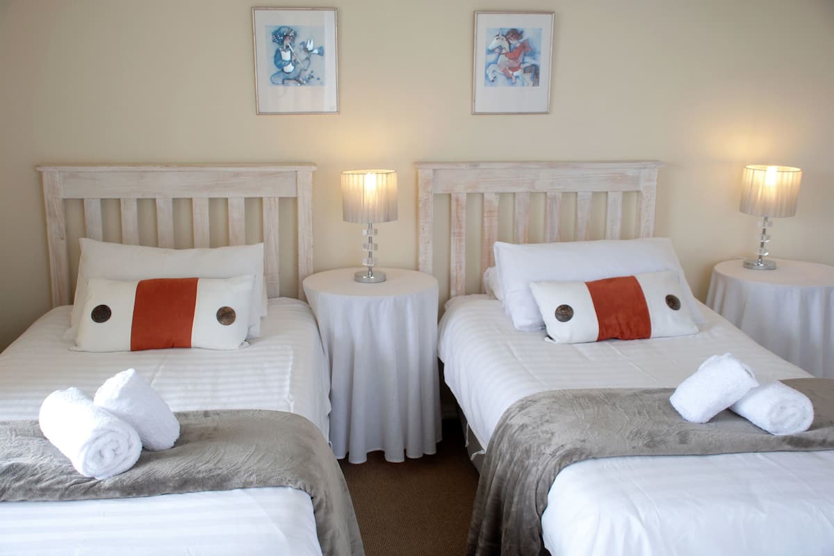 Ein Hotelzimmer mit drei Betten, jeweils mit weißer Bettwäsche und orangefarbenen Akzenten, kleinen runden Tischen mit Lampen und Bildern an den Wänden.