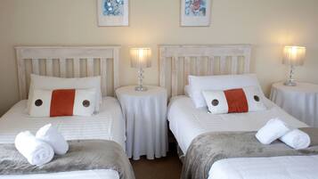 A hotel room with three beds, each with white bedding and orange accents, small round tables with lamps, and artwork on the walls.