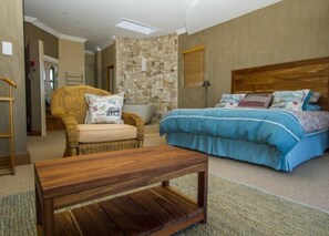 Une chambre avec un lit, une table basse en bois, un fauteuil en osier et un mur en pierre.