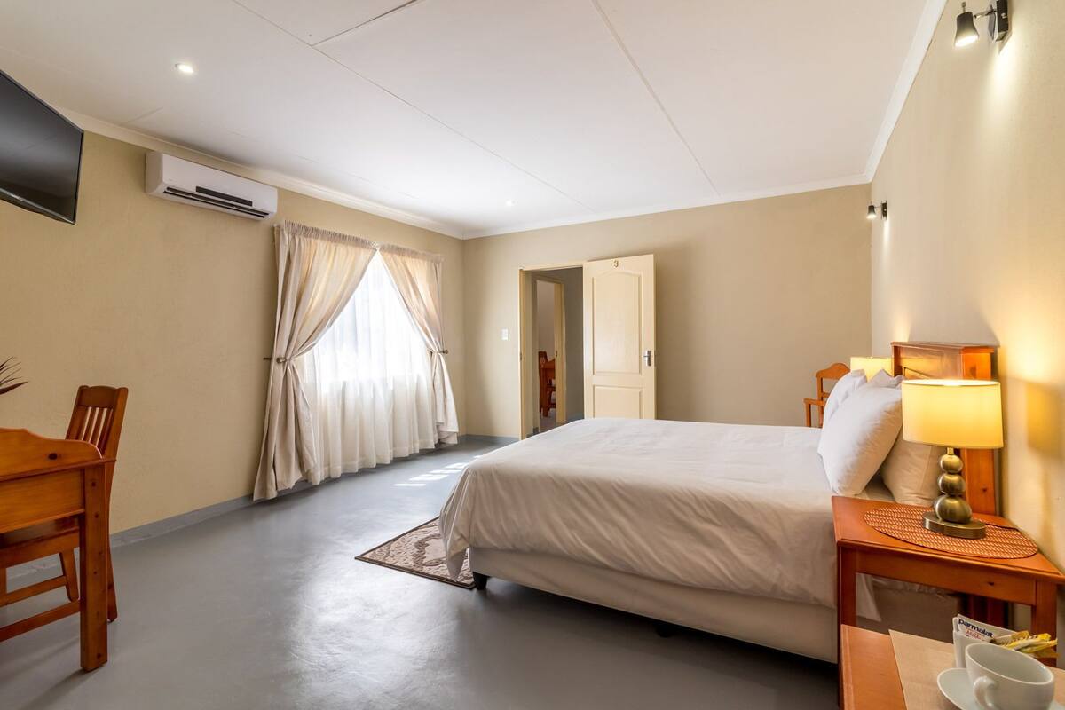 A hotel room with a bed, a desk, a chair, a lamp, and an air conditioning unit.