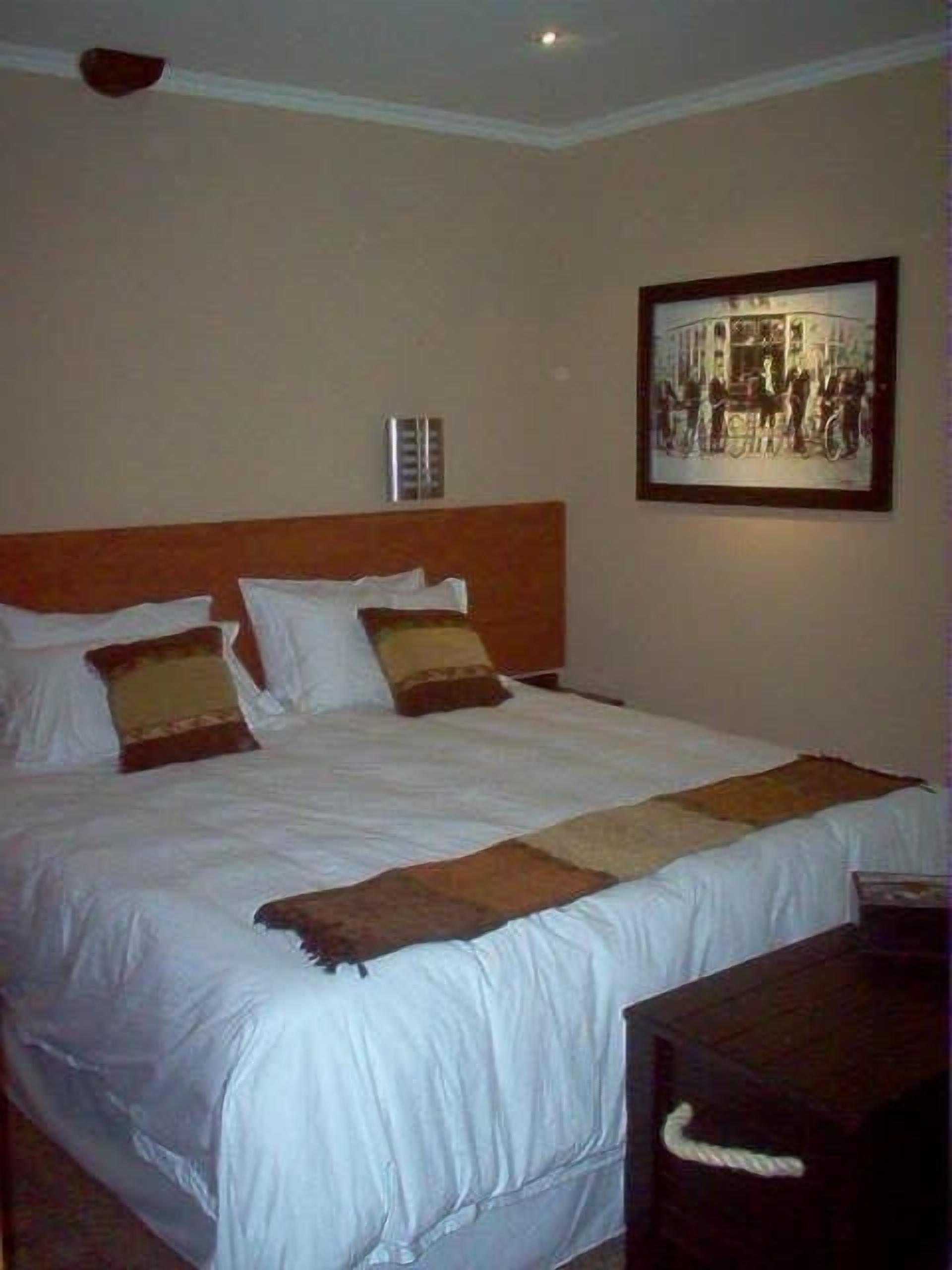Una habitación de hotel con una cama grande, cabecera, mesita de noche, un cuadro enmarcado en la pared y un termostato.