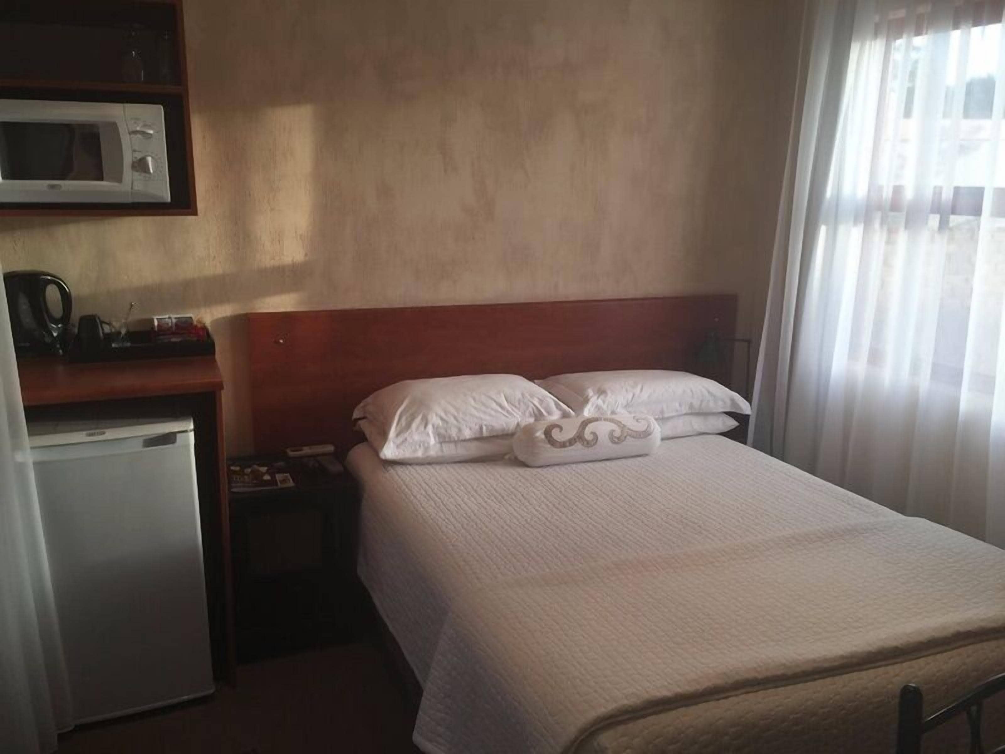 A hotel room with a bed, a microwave, a refrigerator, a nightstand, and a window with curtains.