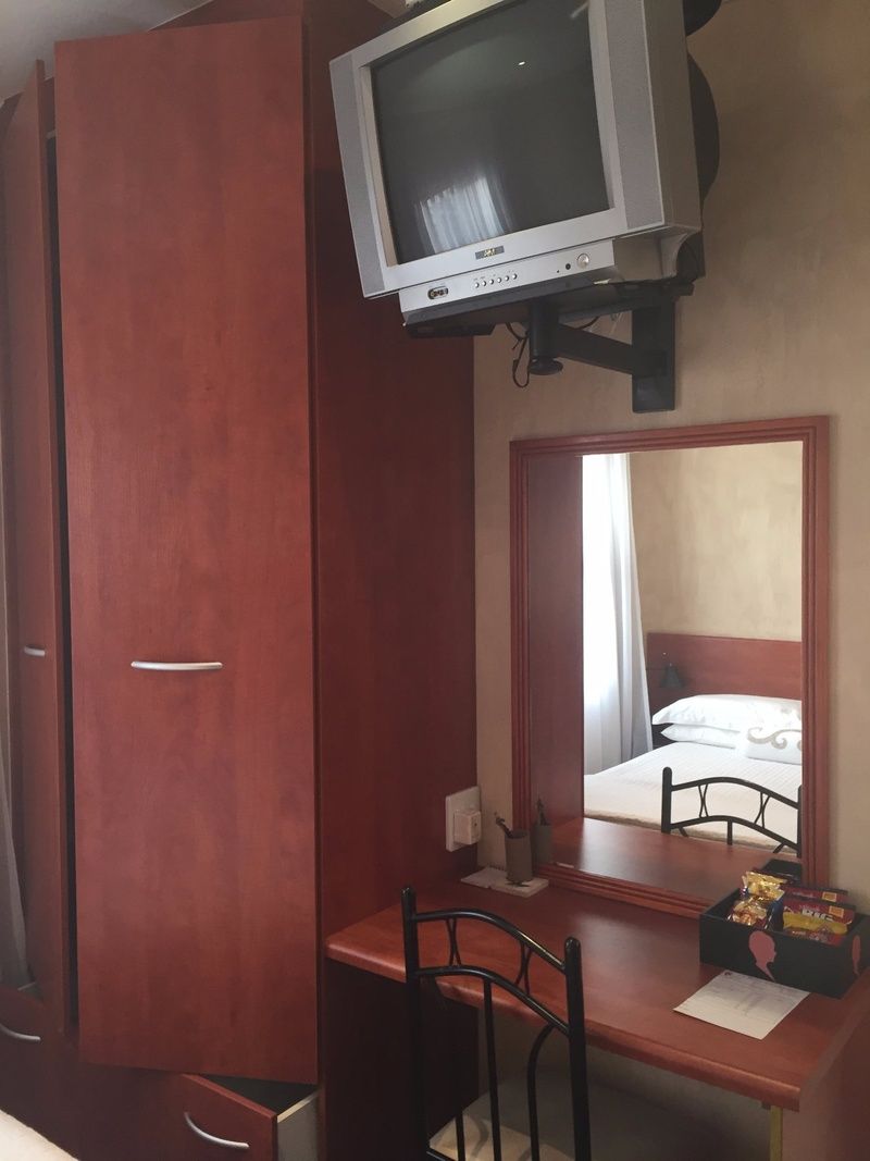 A room with a wooden wardrobe, a desk with a chair, a television mounted on the wall, and a mirror reflecting a bed.