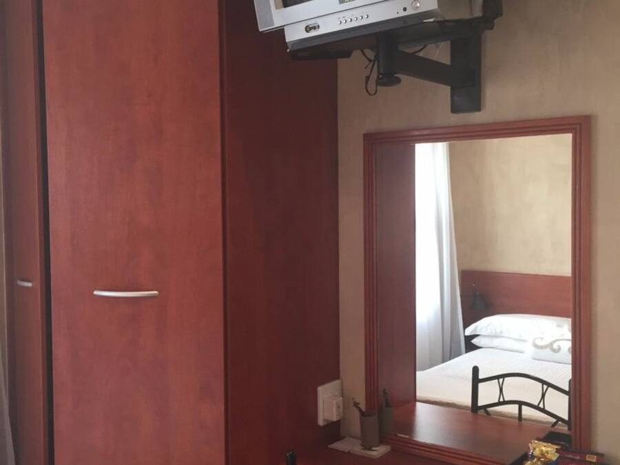 A room with a wooden wardrobe, a desk with a chair, a television mounted on the wall, and a mirror reflecting a bed.