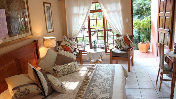 Une chambre bien éclairée, dotée d’un grand lit, de plusieurs oreillers, d’une table de chevet avec une lampe et offrant une vue sur un jardin par la fenêtre.