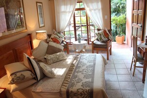 A well-lit bedroom with a large bed, multiple pillows, a bedside table with a lamp, and a view of a garden through a window.
