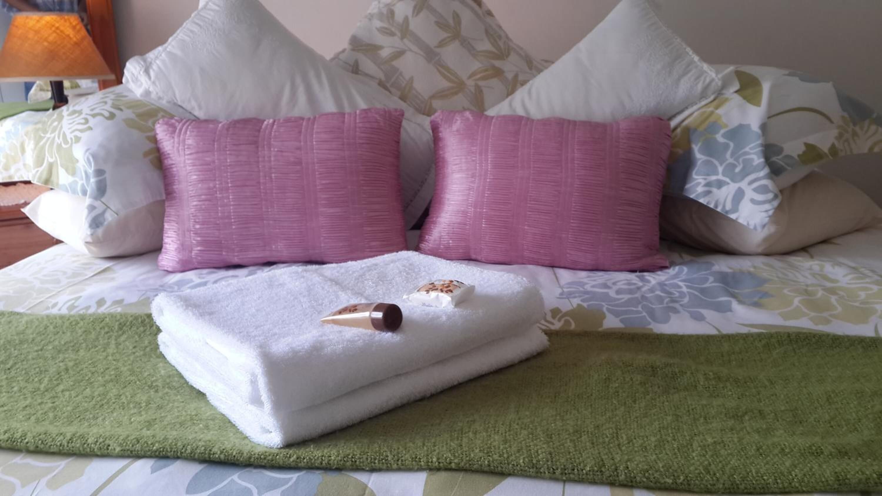 A neatly made bed with a green blanket, two pink pillows, and a white towel with a bottle on it.
