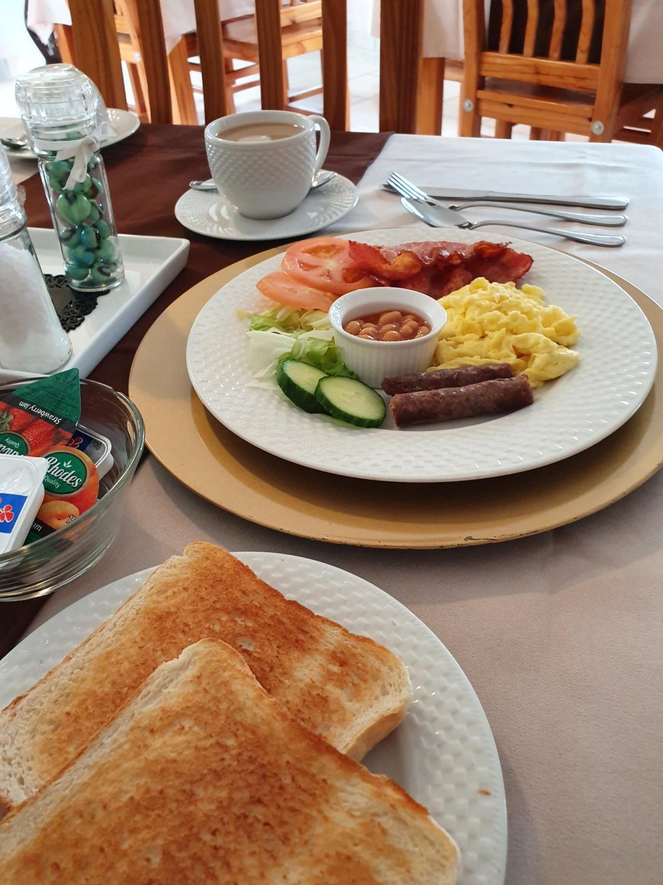 A breakfast setting with toast, scrambled eggs, bacon, tomatoes, cucumbers, and beans on a plate, along with a cup of coffee and a glass of water.