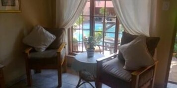 A room with two wooden chairs and a small round table, a window with curtains, and a view of a swimming pool.