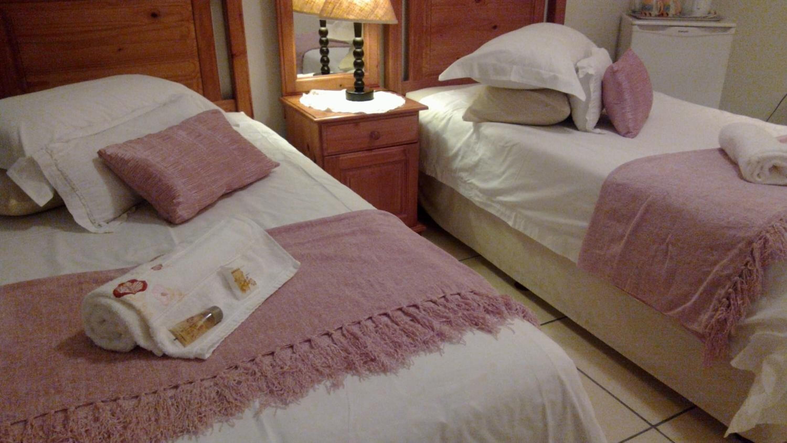 Ein Hotelzimmer mit zwei Betten, jeweils mit weißer Bettwäsche und rosafarbenen Akzenten. Zwischen den Betten steht ein Nachttisch aus Holz mit einer Lampe.