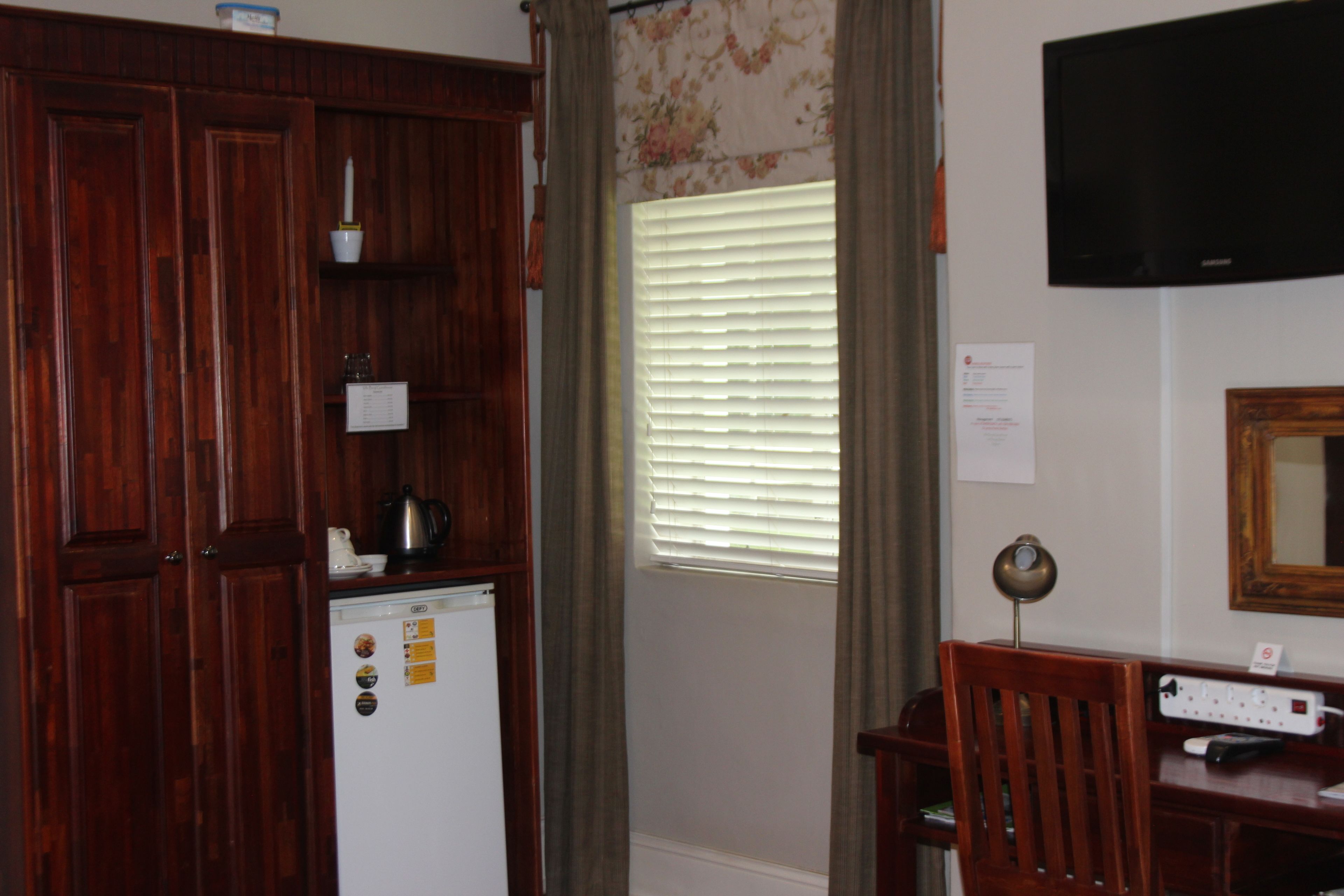 A room with a wooden wardrobe, a white refrigerator, a window with blinds, a desk with a lamp, and a flat-screen TV mounted on the wall.
