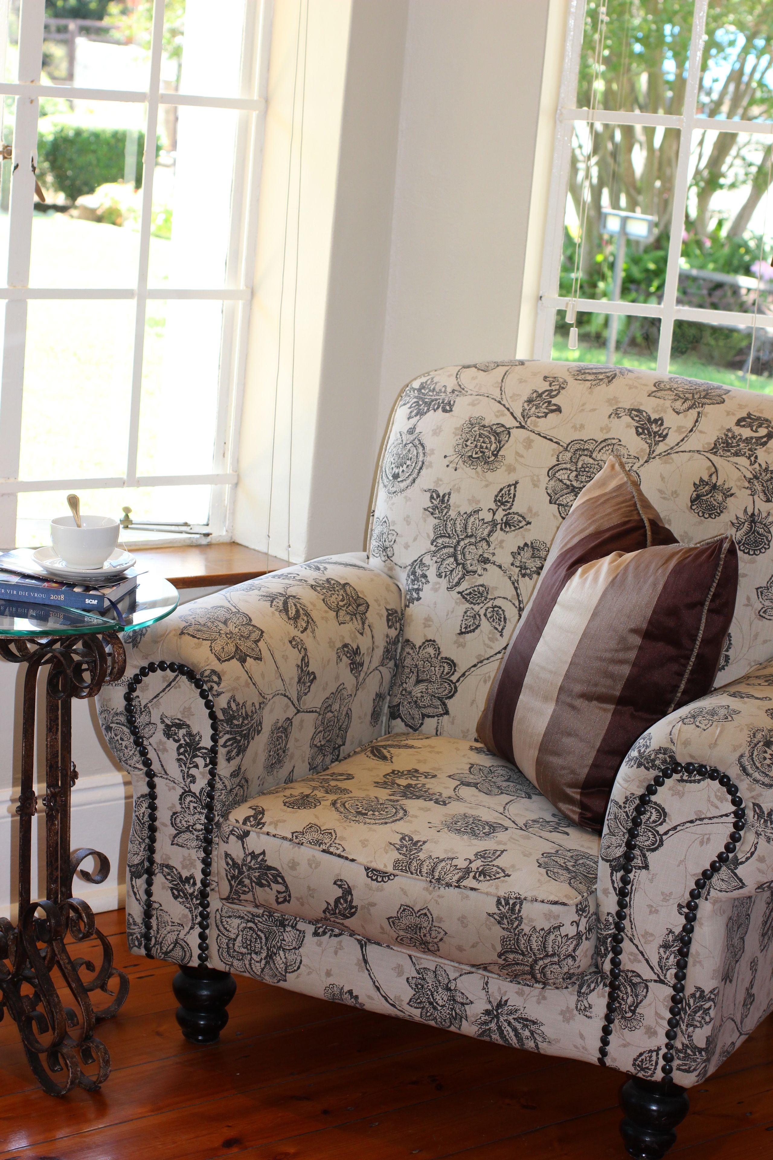 Un sillón con estampado floral y un cojín, una mesita de cristal con un cuenco y libros, y una ventana con vistas a zonas verdes.
