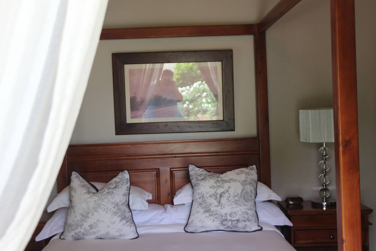 A bedroom with a wooden headboard, a bed with floral-patterned pillows, a bedside table with a lamp, and a framed picture on the wall.