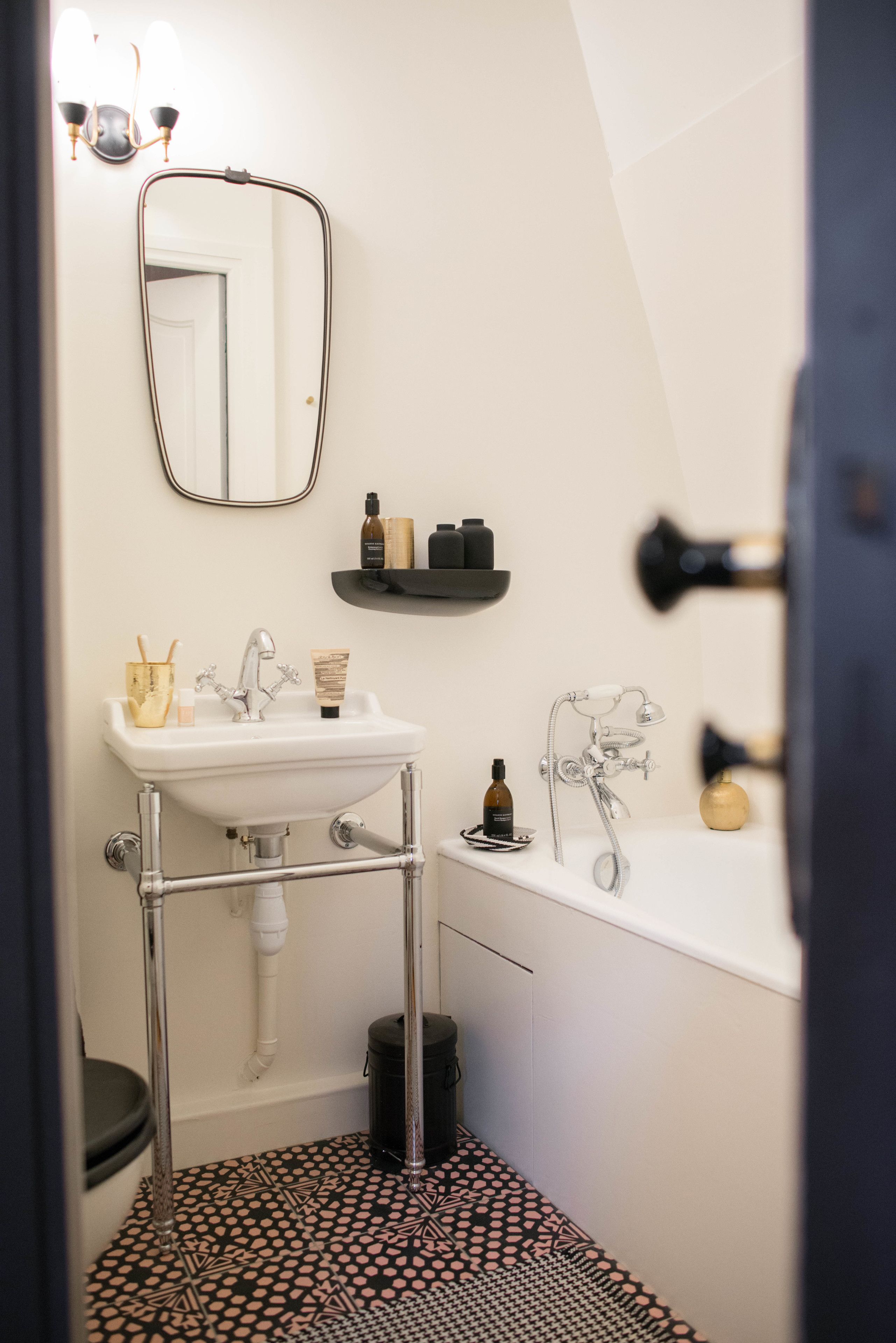 A bathroom with a white sink, mirror, and bathtub. There are toiletries on the sink and a black trash can.