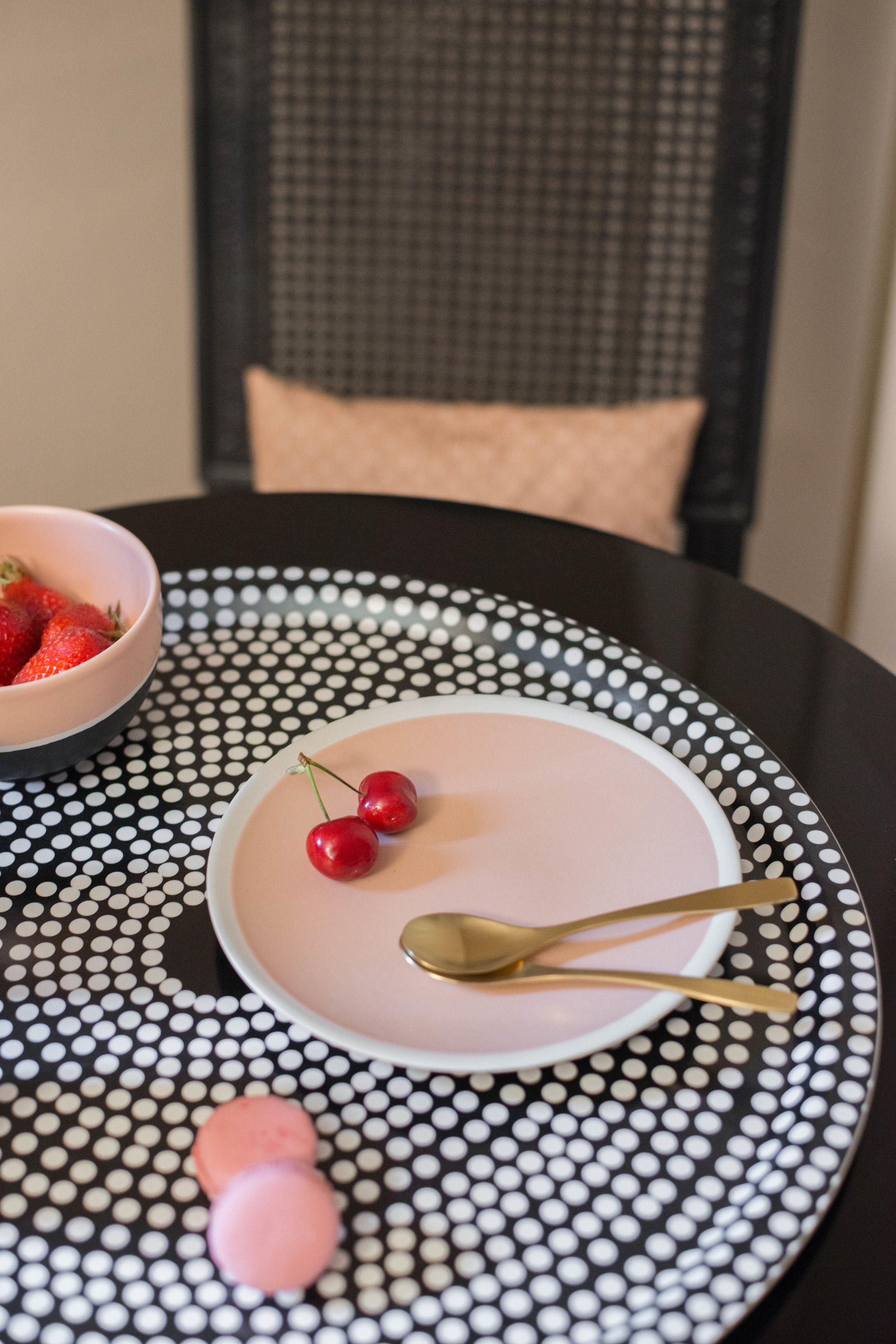 A table with a polka dot tablecloth, a pink plate with cherries and a spoon, and a bowl of strawberries.