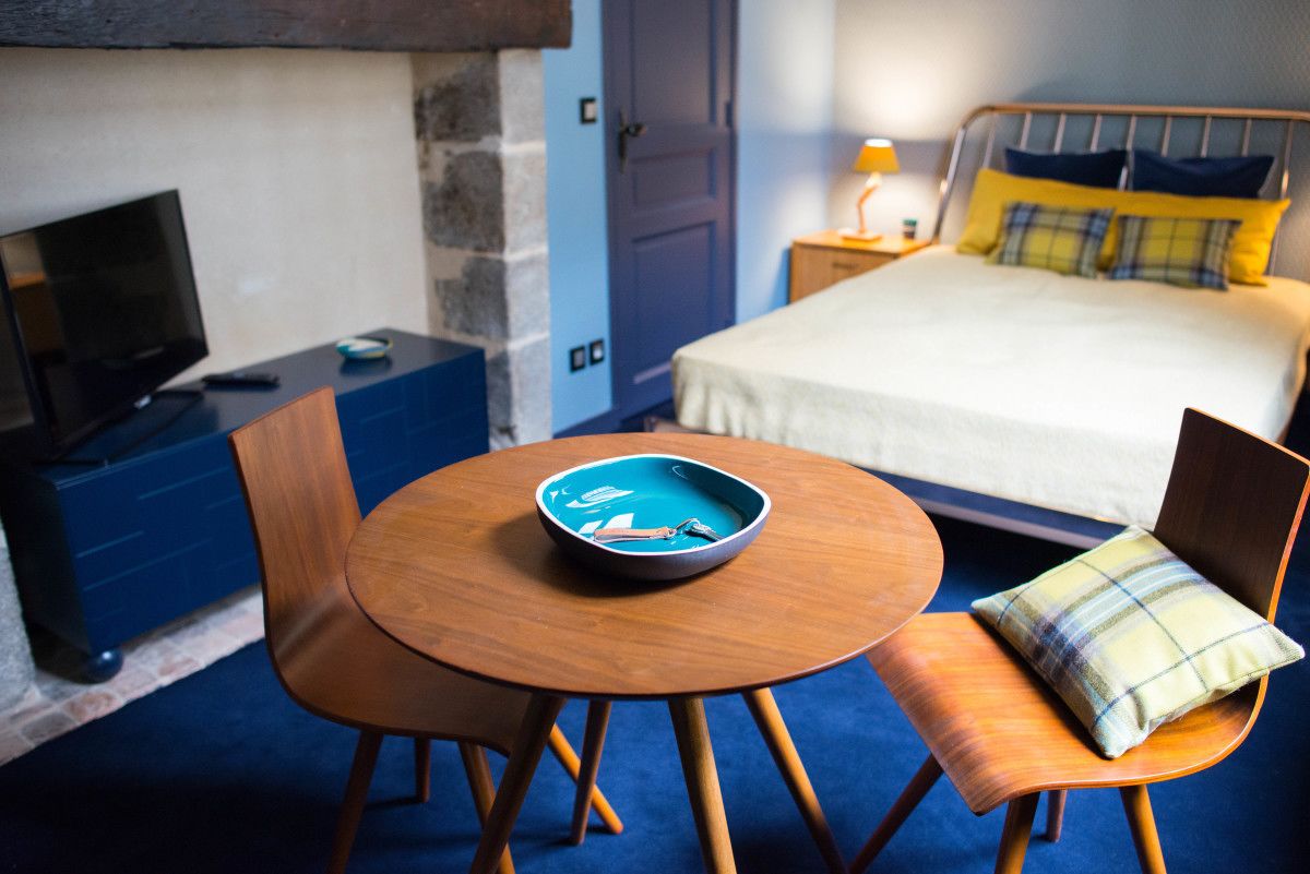 Une chambre d’hôtel avec un lit, une télévision, une table ronde en bois sur laquelle se trouve un bol, et une chaise avec un coussin à carreaux.