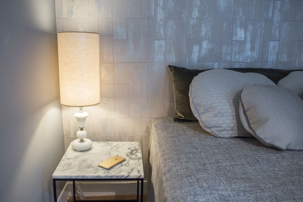 A neatly made bed with a grey blanket and pillows, a white marble nightstand with a lamp, and a smartphone on the nightstand.