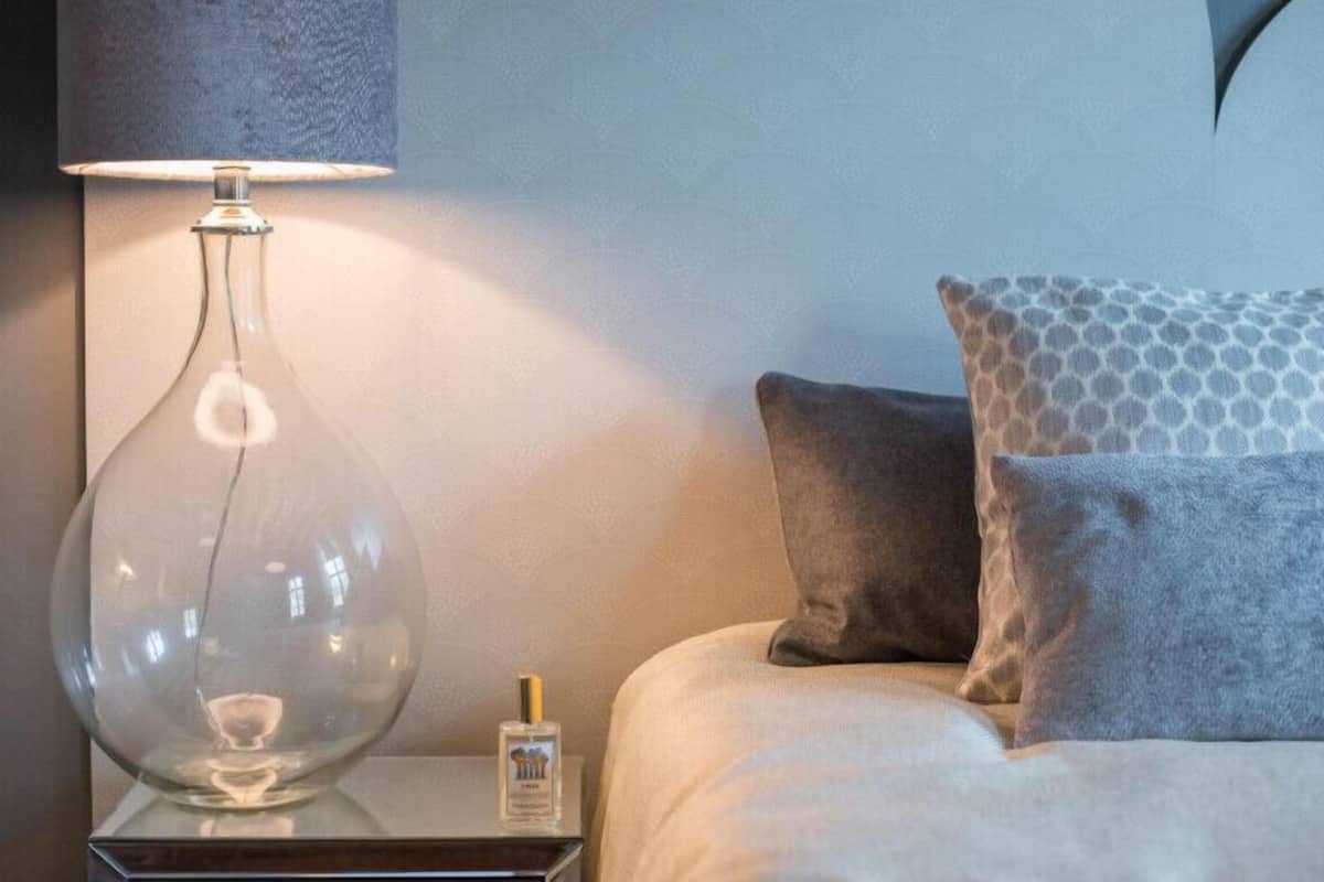 A modern bedroom with a clear glass lamp on a bedside table, a bottle of hand lotion, and two patterned pillows on a bed.
