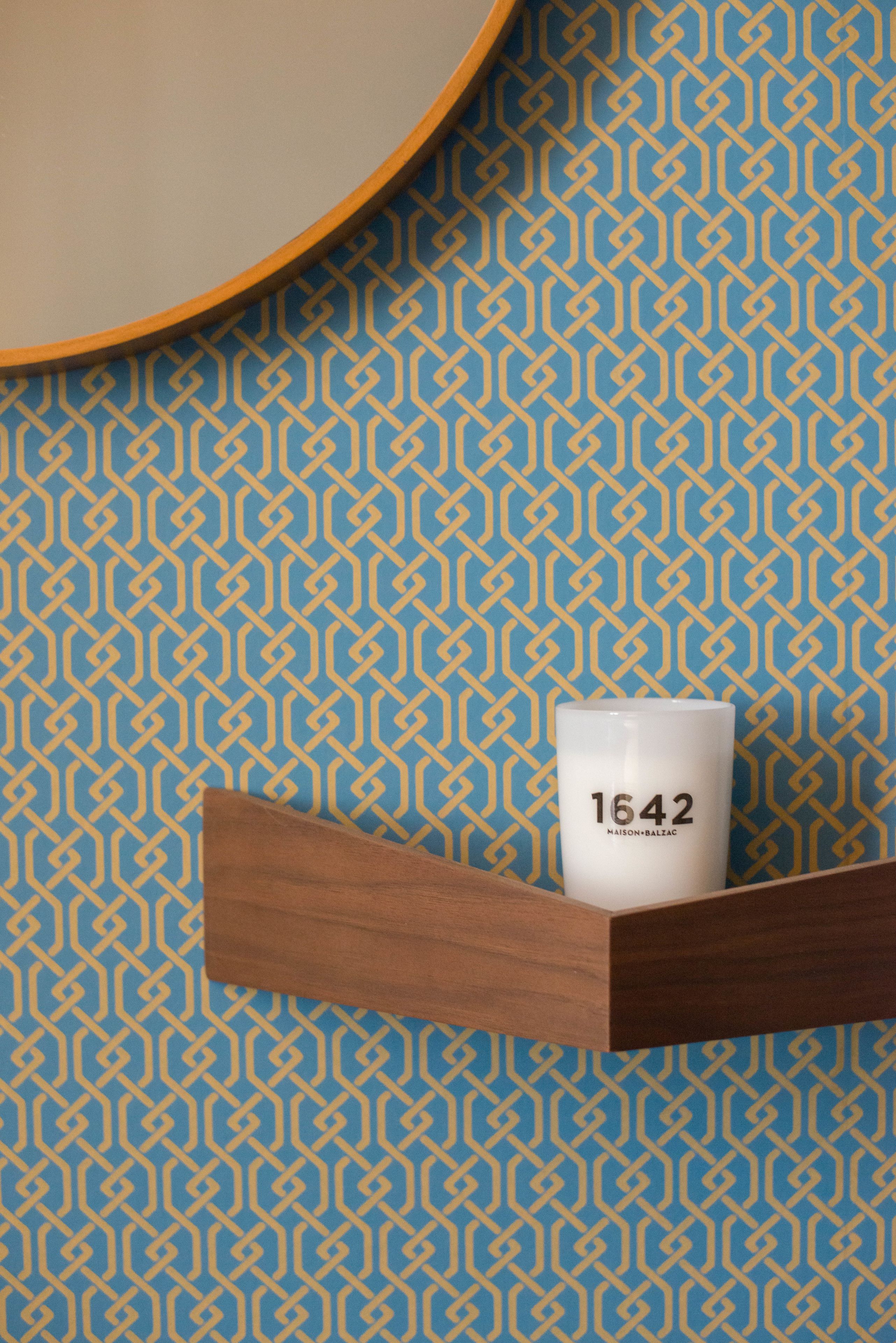 Uma caneca branca com a inscrição '1642', apoiada numa prateleira de madeira em frente a uma parede azul estampada.