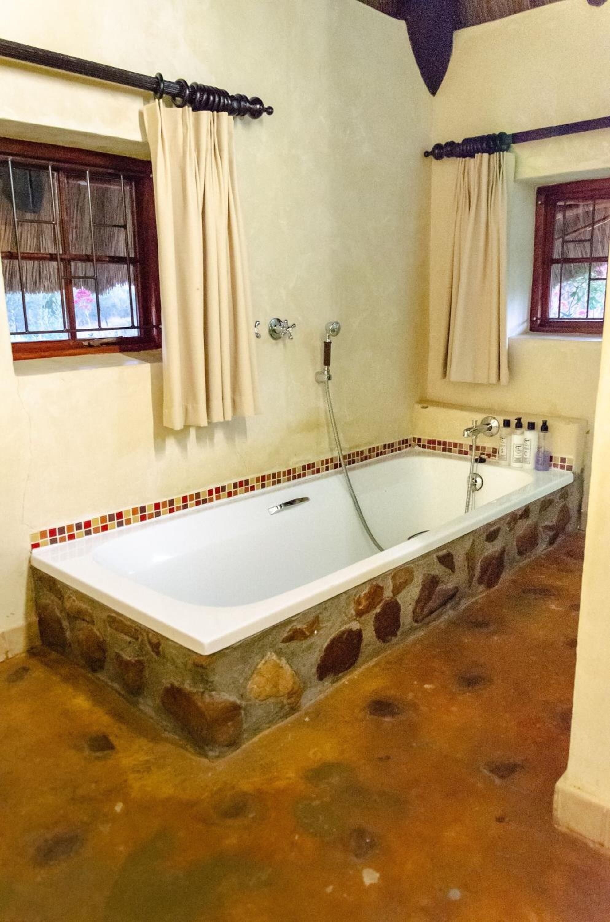 Un baño con una bañera de piedra, dos ventanas con cortinas y piso de baldosas.