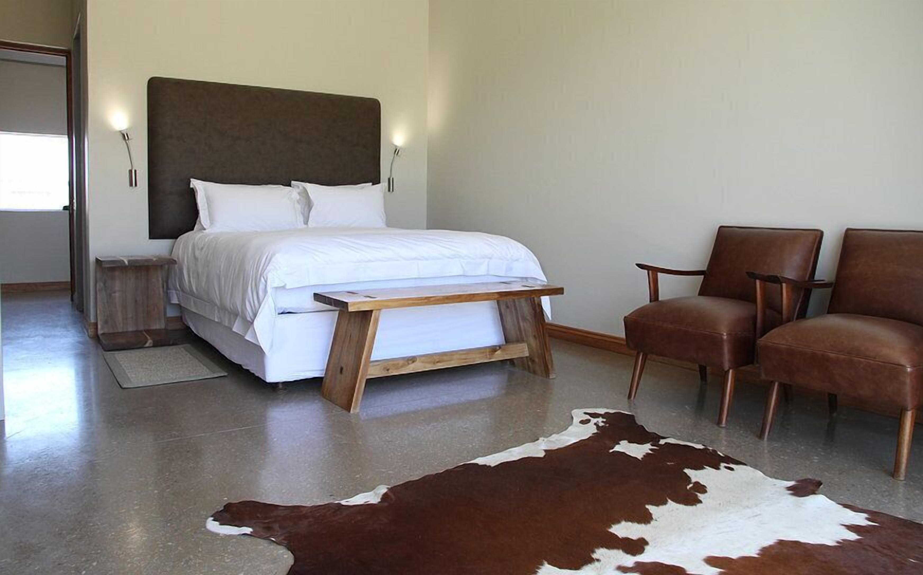 Une chambre à coucher moderne avec un lit, deux chaises, une table de chevet en bois, un tapis en peau de vache et une commode.