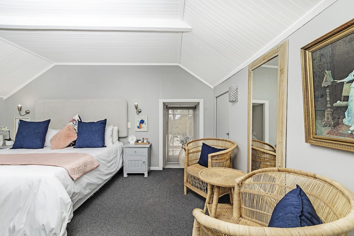 Une chambre avec un grand lit, une table de chevet, une chaise en osier, un miroir et une porte donnant sur une autre pièce.