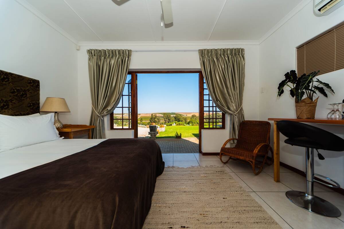 A bedroom with a bed, a desk, a chair, and a view of a green landscape through a window.