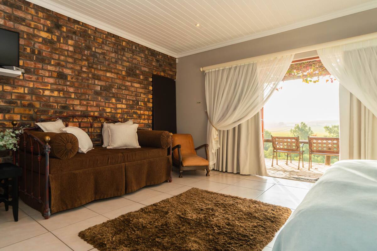 A room with a brick wall, a brown sofa, a wooden chair, a large rug, and a view through a window with curtains.