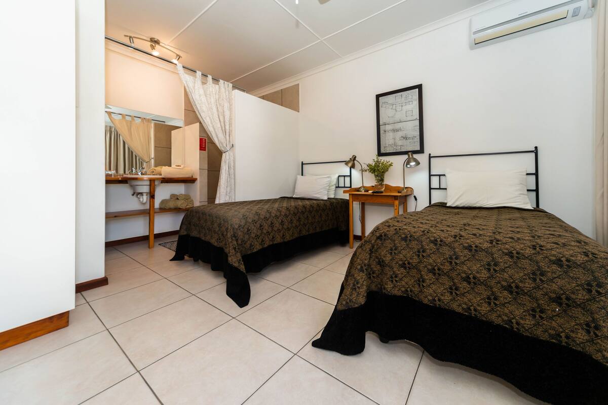 A bedroom with two beds, a bedside table with a lamp, a mirror, and a window with a curtain.