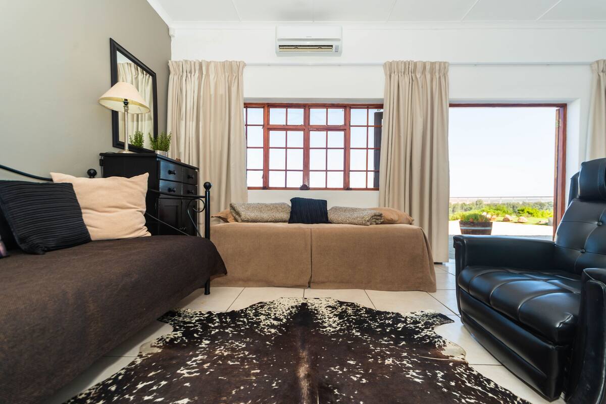 A modern living room with a sofa, a black leather chair, a large window with curtains, and a ceiling fan.