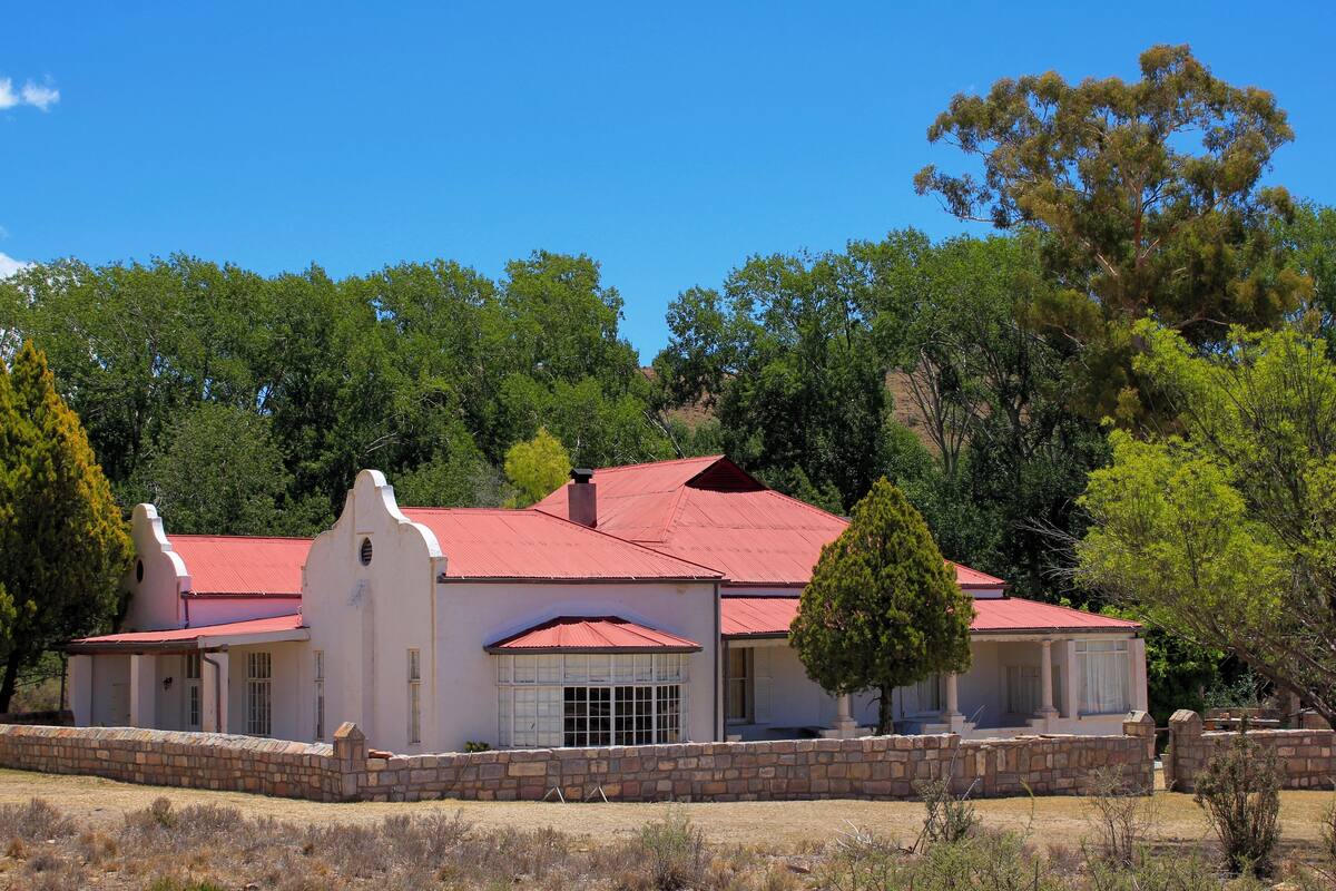 A two-story building with a red roof and white walls, surrounded by trees and a stone wall.