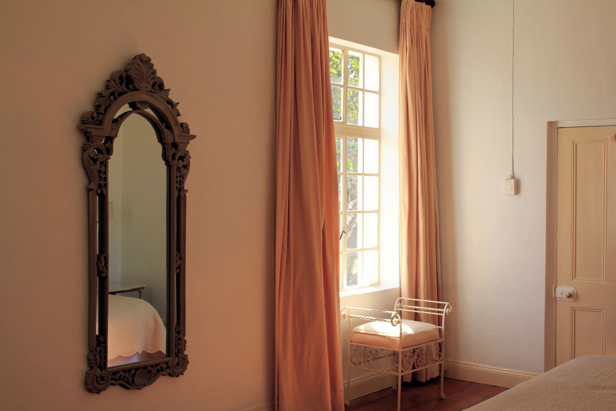 A bedroom with a large ornate mirror, a window with curtains, a metal bedside table, and a door.