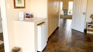 A kitchen with a white countertop, a coffee maker, a kettle, and a basket. There is a wooden cabinet with drawers and a door. The floor is tiled, and there is a door leading to another room. - Marina Martinique B and B (Jeffreys Bay)