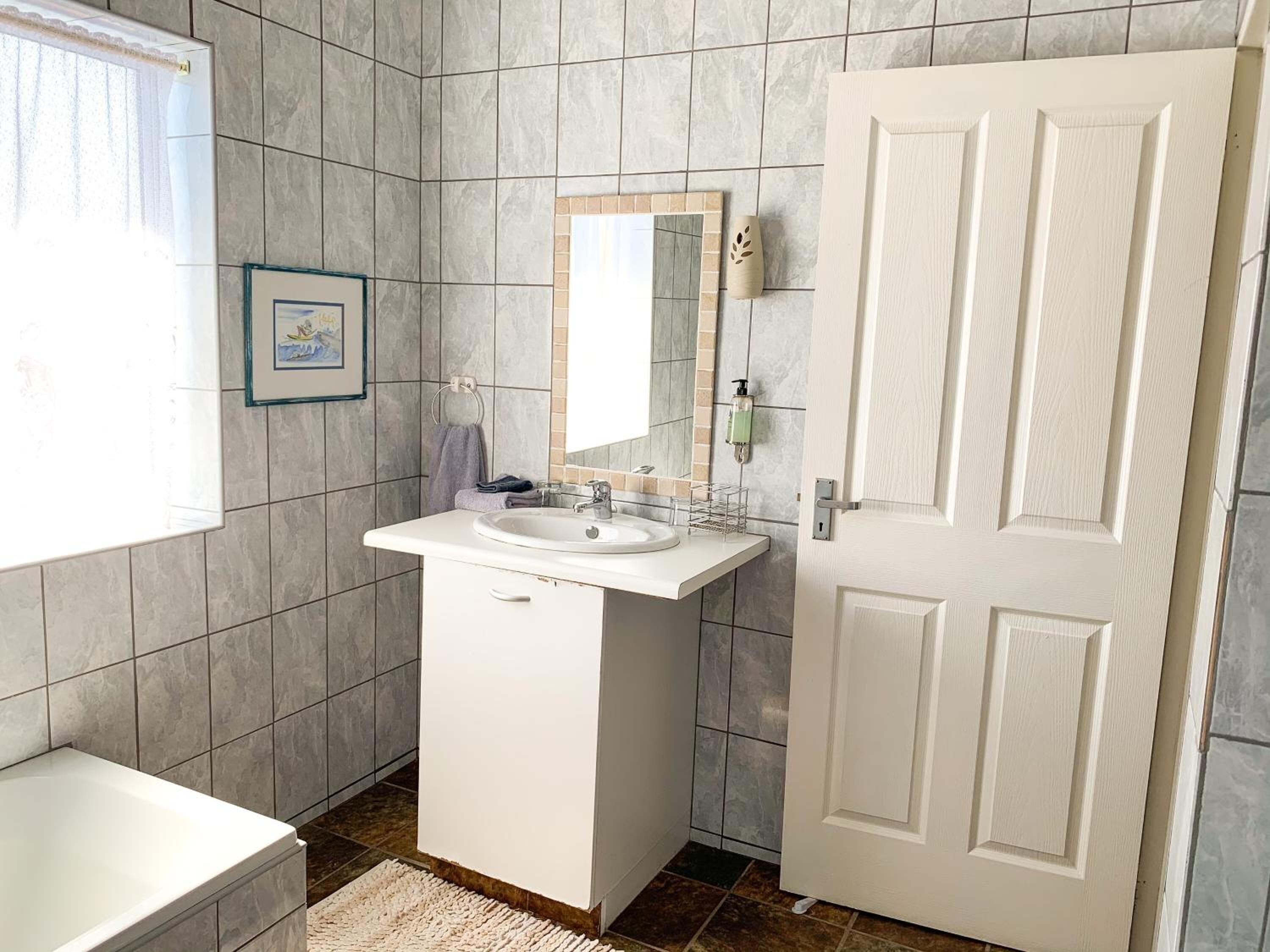 A bathroom with a white sink, mirror, and door, tiled walls, and a window.
