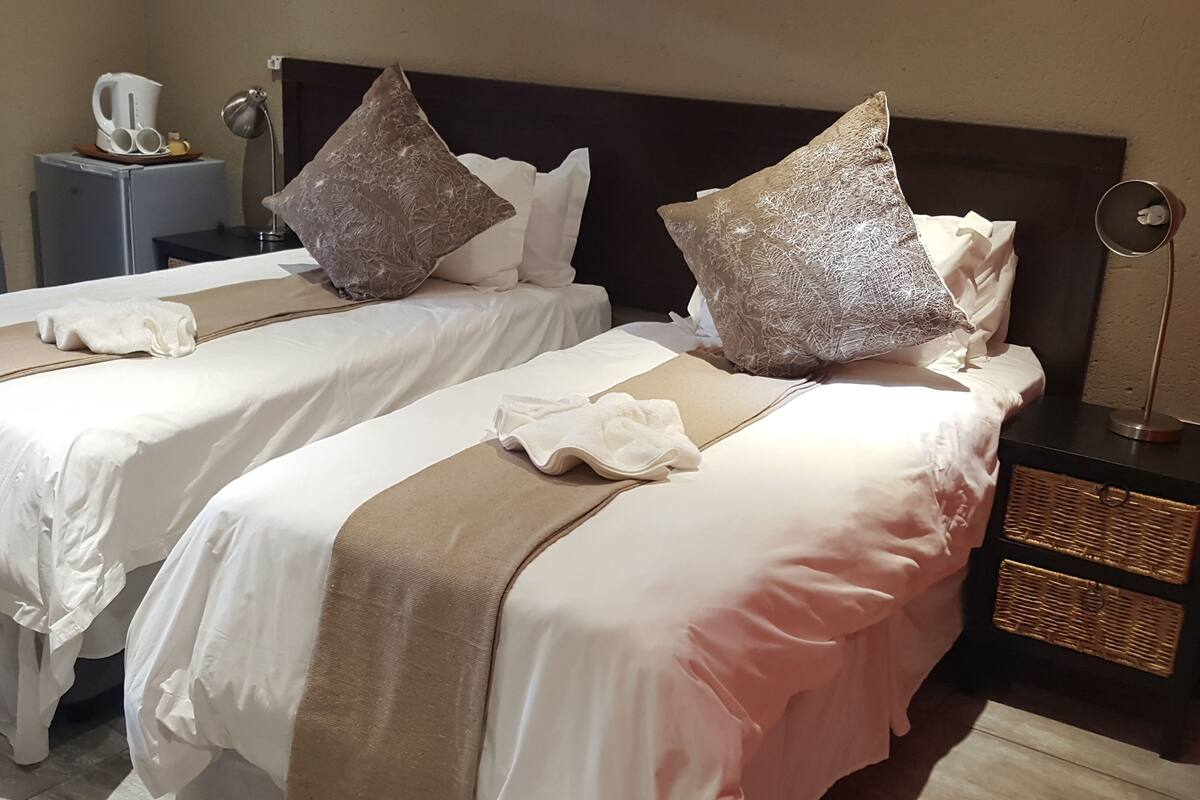 A hotel room with two beds, each with white linens and decorative pillows. There is a bedside table with a lamp and a small basket.