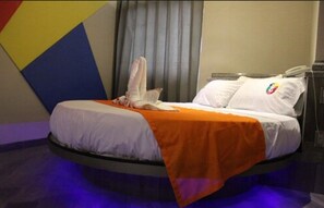 A bed with a white mattress and orange blanket, a white pillow with a colorful logo, and a telephone on the side.
