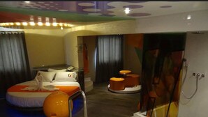 A modern hotel room with a bed, a bathroom with a glass shower enclosure, and a small seating area with stools.
