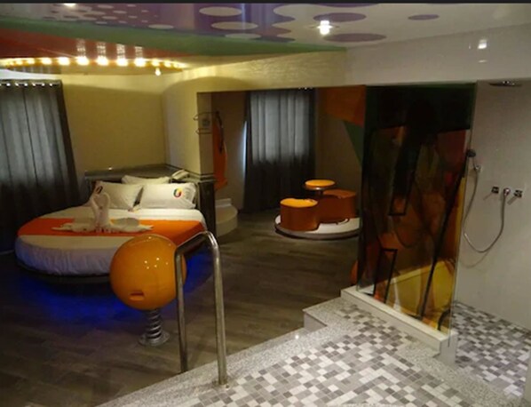 A modern hotel room with a bed, a bathroom with a glass shower enclosure, and a small seating area with stools.