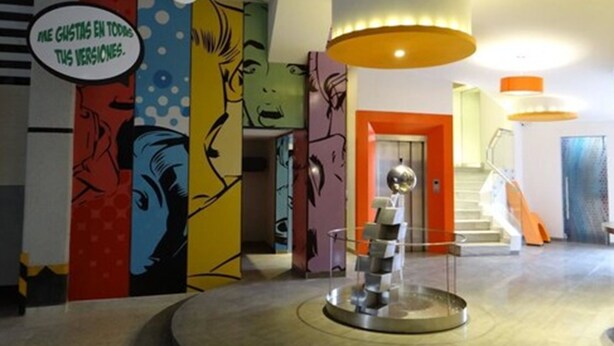 A lobby with colorful comic-style murals on the walls, a central metallic sculpture, and a staircase with orange accents.