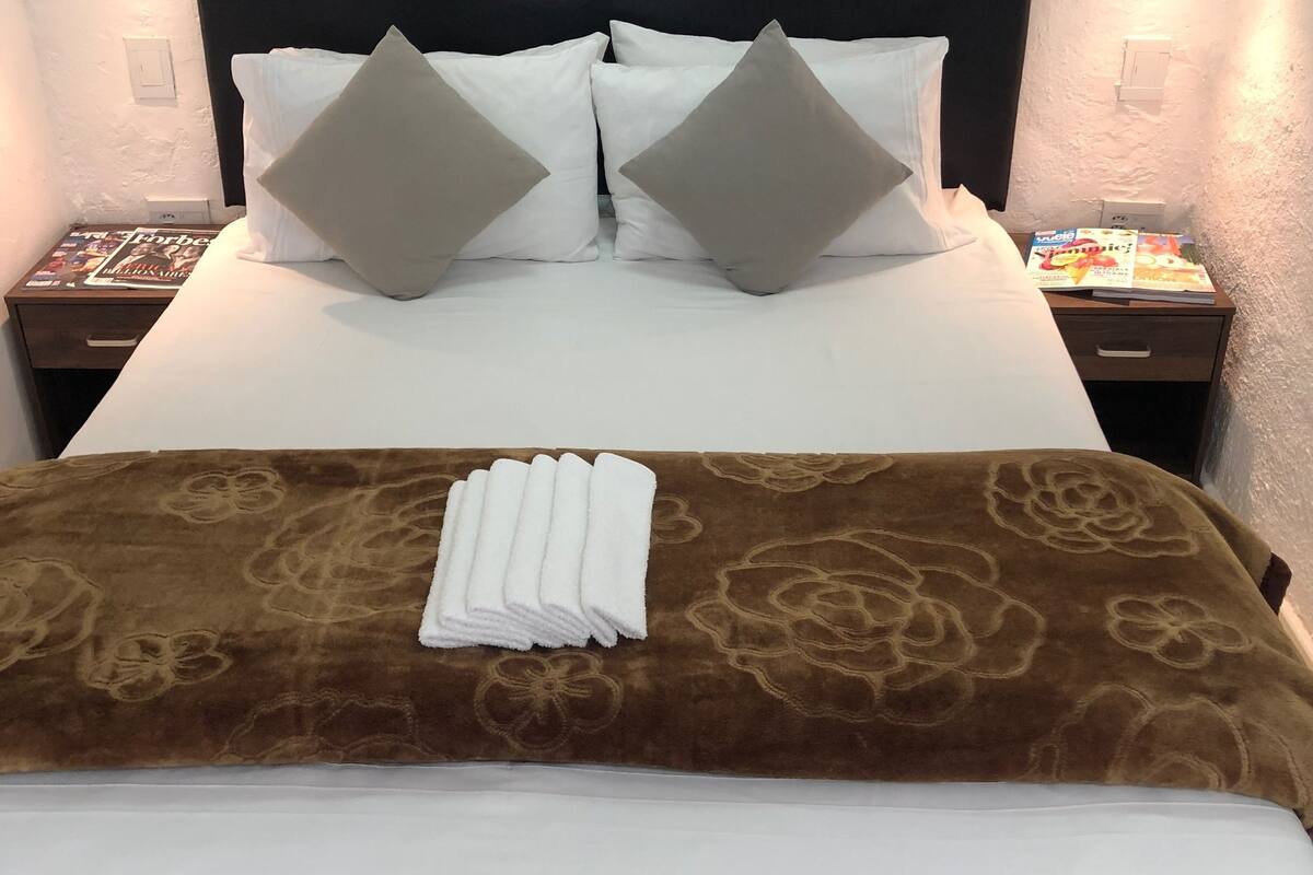 Un lit bien fait, recouvert d’une couverture à motifs floraux marron, avec des oreillers blancs et une serviette blanche pliée. Il y a deux tables de chevet, chacune avec une lampe, et sur l’une d’elles, on peut voir un petit magazine.