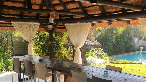 A covered outdoor dining area with a wooden table and chairs, a pool in the background, and a thatched roof overhead. - Muco Guest House (Sandton)