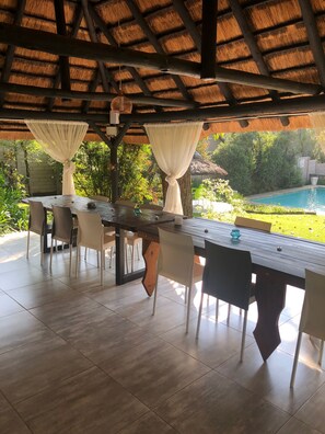 A covered outdoor dining area with a wooden table and chairs, a pool in the background, and a thatched roof overhead. - Muco Guest House (Sandton)