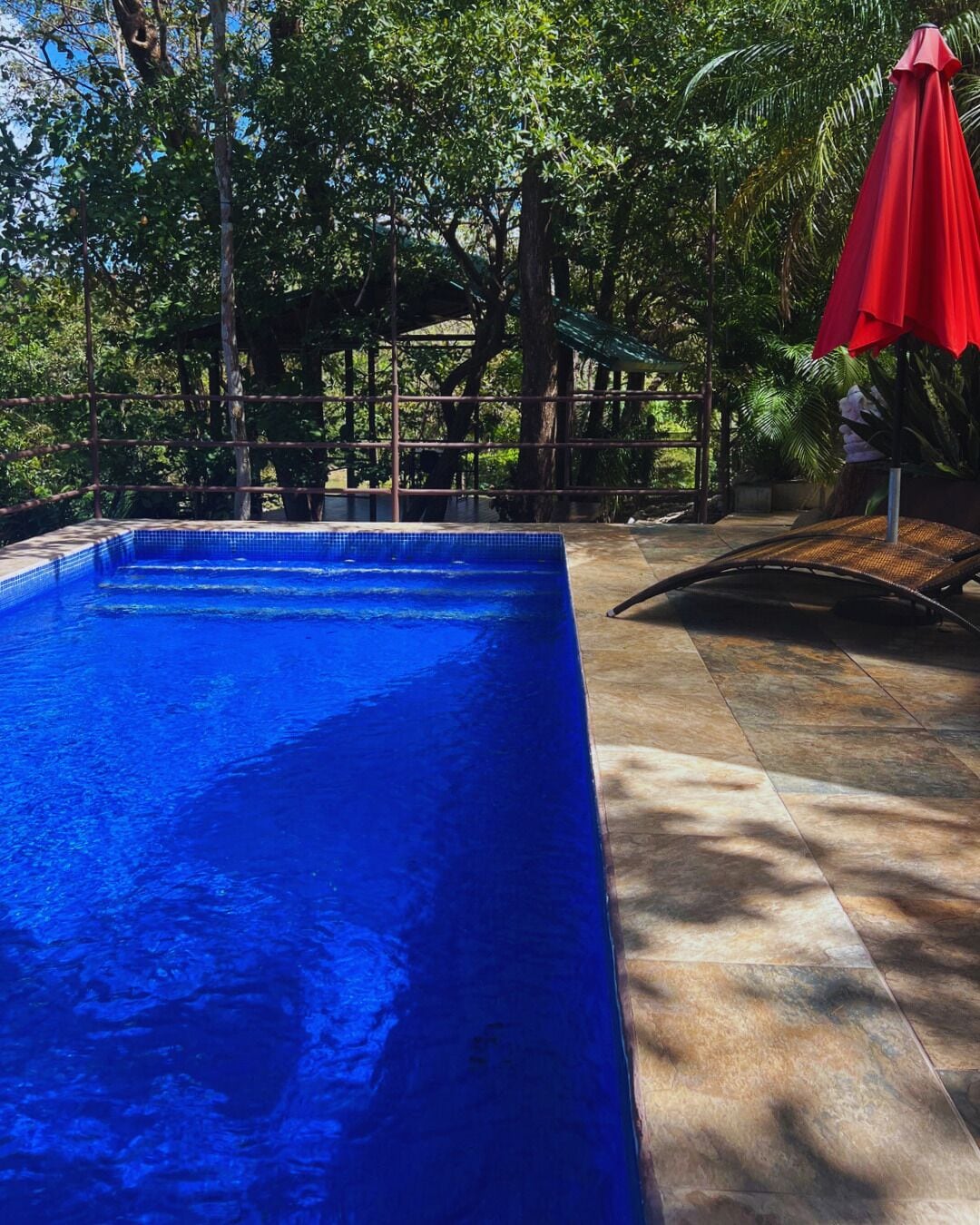 A rectangular swimming pool with clear blue water, surrounded by a tiled deck and a red umbrella.