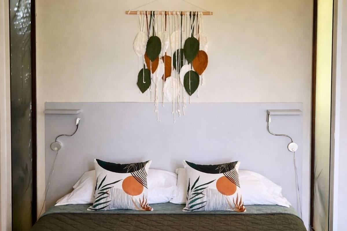 A bed with a dark quilted bedspread, two pillows with leaf patterns, a wall hanging with hanging elements, and a rolled towel on the bed.