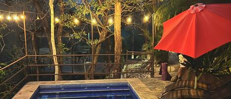 Una piscina rectangular con borde de azulejos, rodeada de árboles y un paraguas rojo.