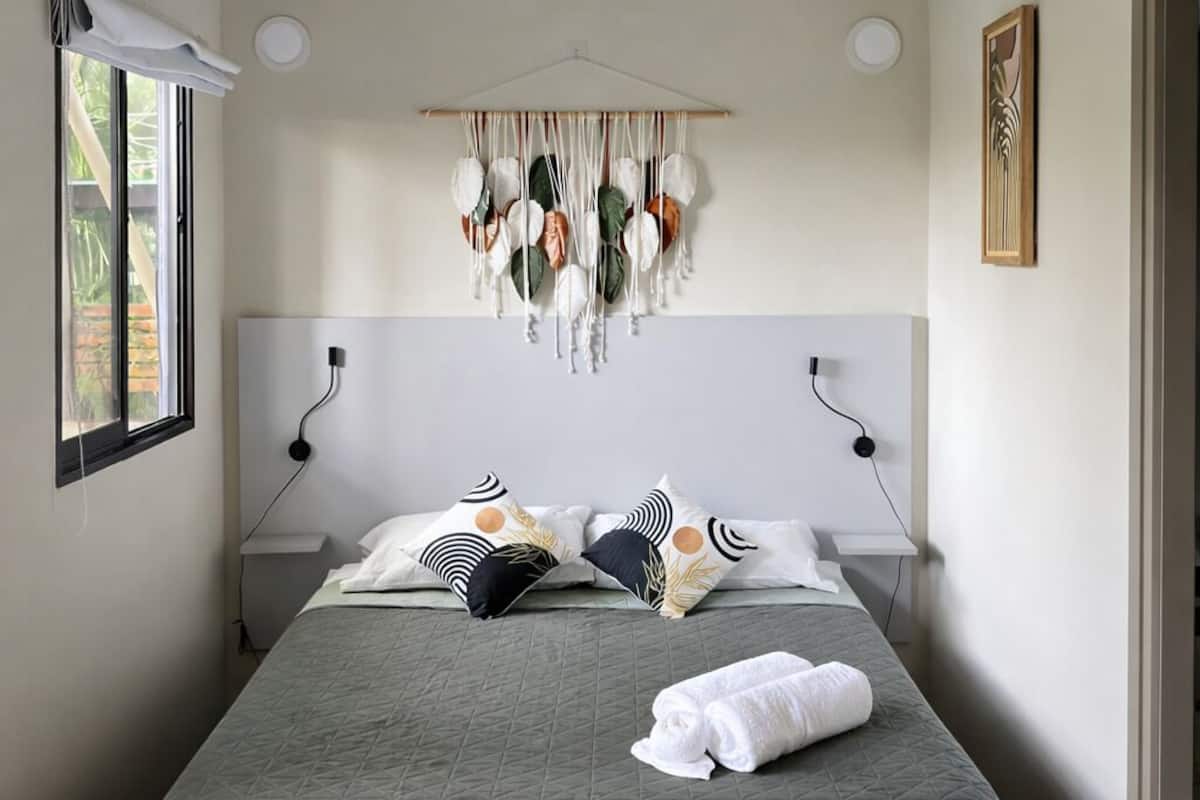 A bed with a quilted bedspread, a hanging macrame wall decoration, a ceiling fan, and a painting on the wall.