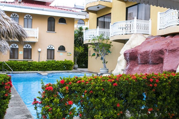 Zona de piscina con una formación rocosa, rodeada de vegetación y un edificio con balcones.