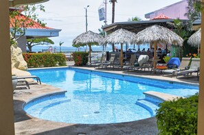 Piscina con sillones de playa y sombrillas, rodeada de vegetación y vista al océano.