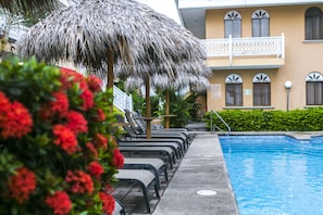A pool area with lounge chairs and a thatched umbrella, a building with balconies, and a sign indicating a swimming area. - Hotel Alamar (Puntarenas)